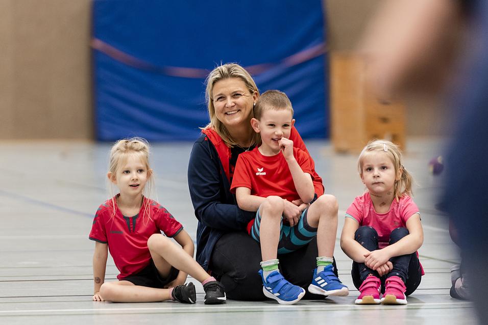 Eine Trainerin sitzt mit drei Kindern in Sportkleidung auf dem Boden in einer Sporthalle.